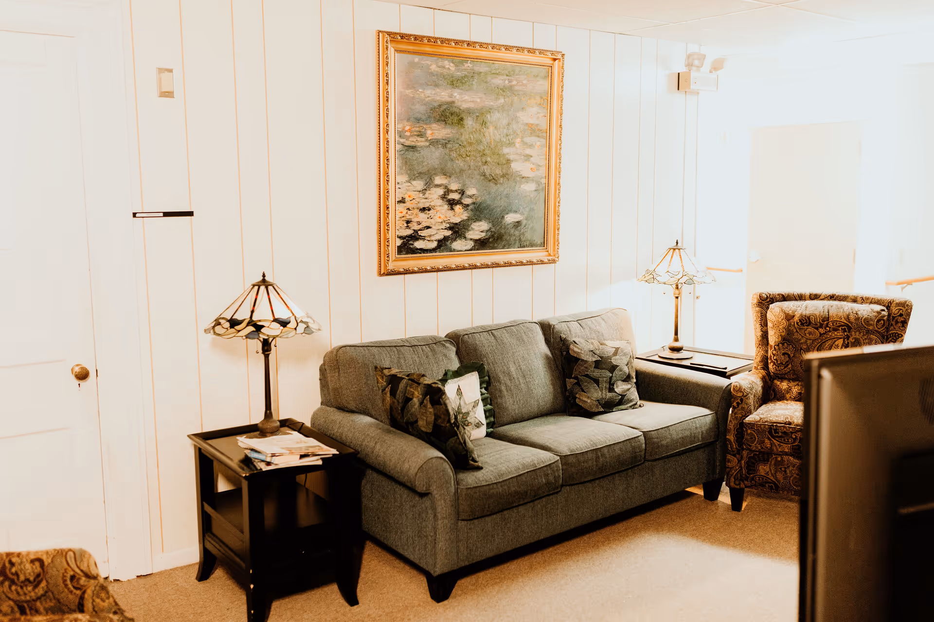 A cozy living room area with a gray sofa adorned with patterned pillows, a patterned armchair, two side tables each with a decorative lamp, and a framed painting of water lilies on the wall behind the sofa.