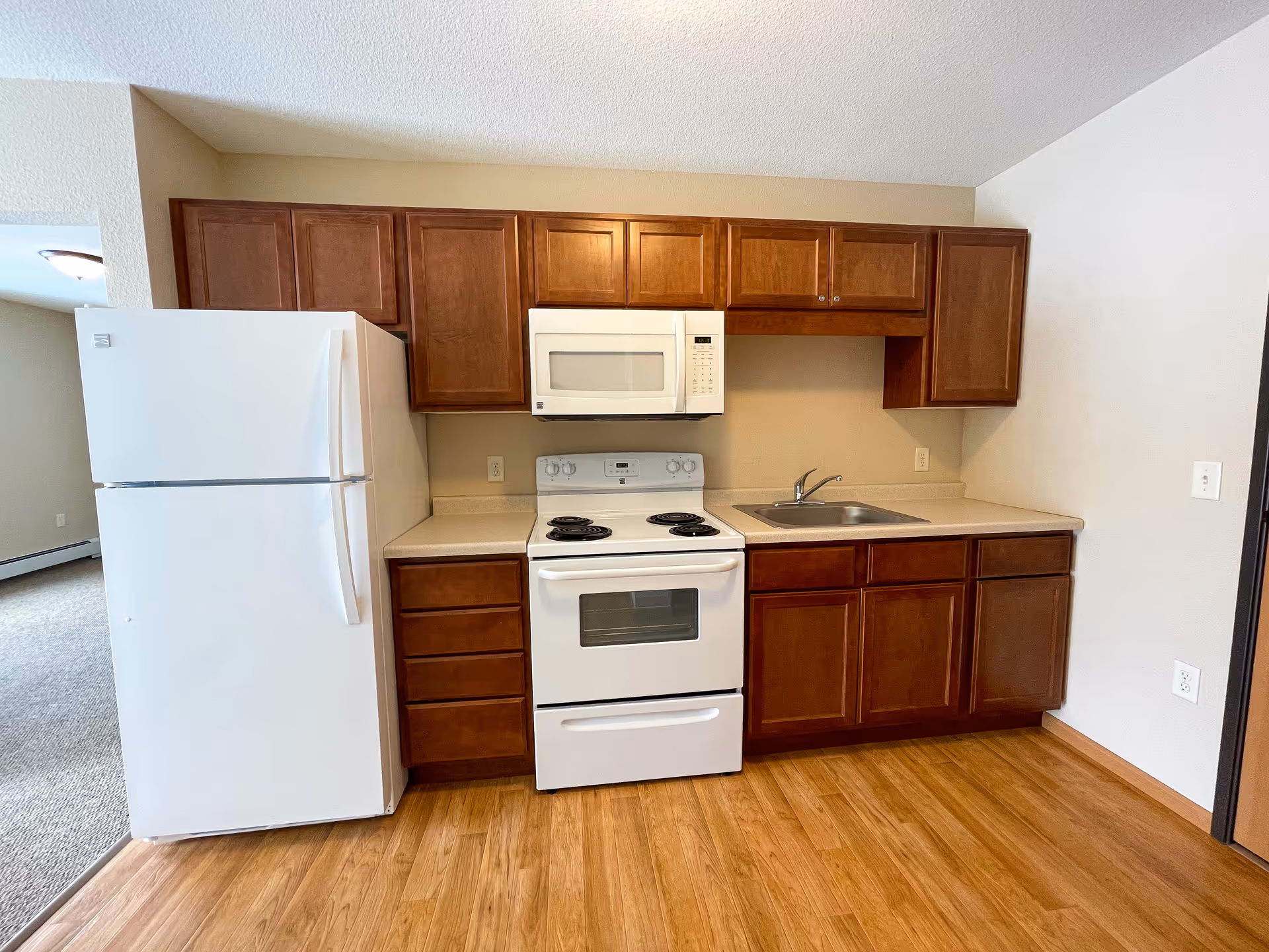 A small kitchen area with wooden cabinets, a white refrigerator, a white electric stove with oven, a white microwave above the stove, a sink with a faucet, and a light-colored countertop. The floor is wooden and the walls are painted beige.