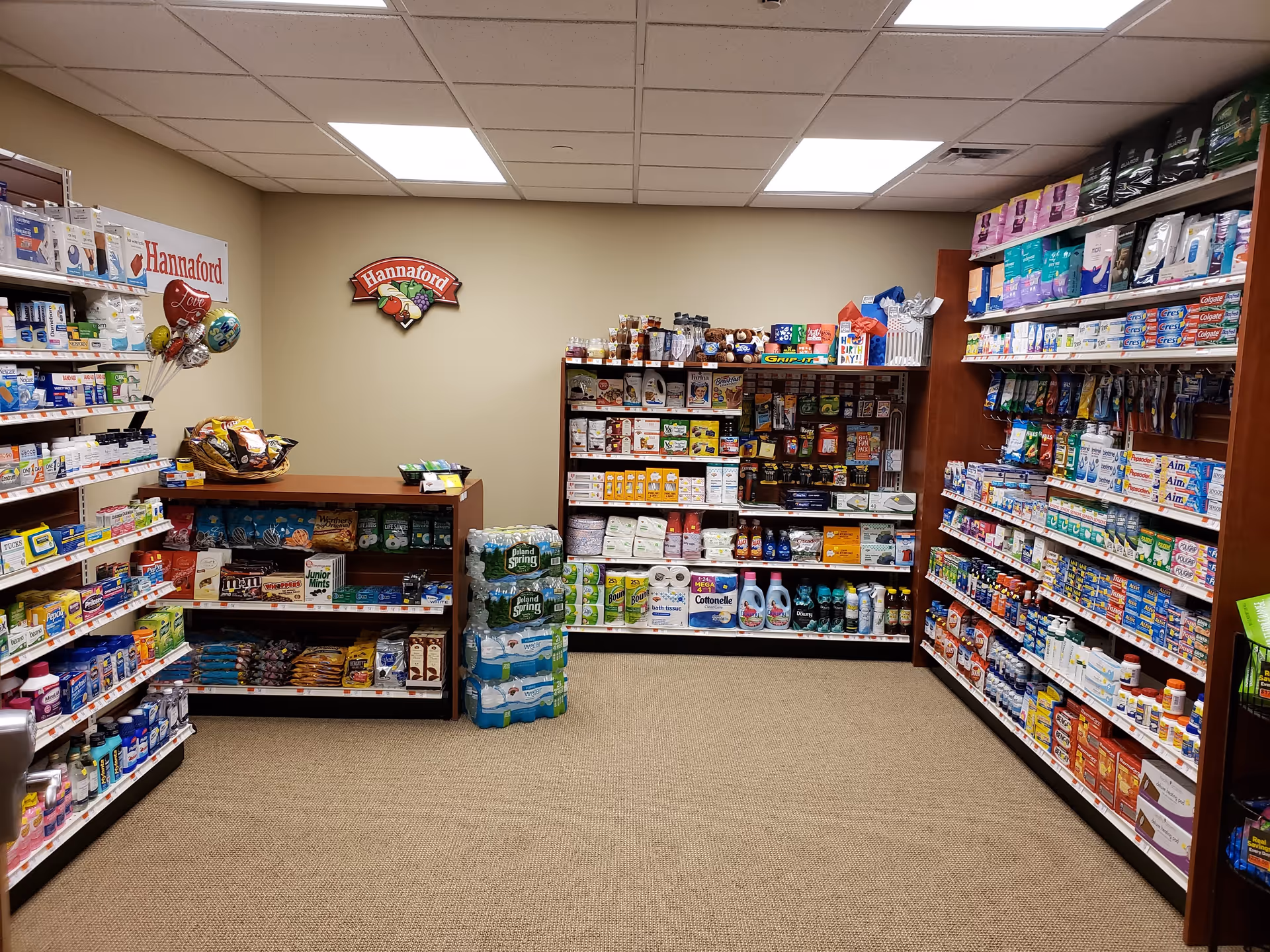 Interior view of a small convenience store or market area with shelves stocked with various grocery and household items including snacks, bottled water, cleaning supplies, and personal care products. The walls are beige, and there is a ceiling with fluorescent lighting. A Hannaford sign is mounted on the back wall.