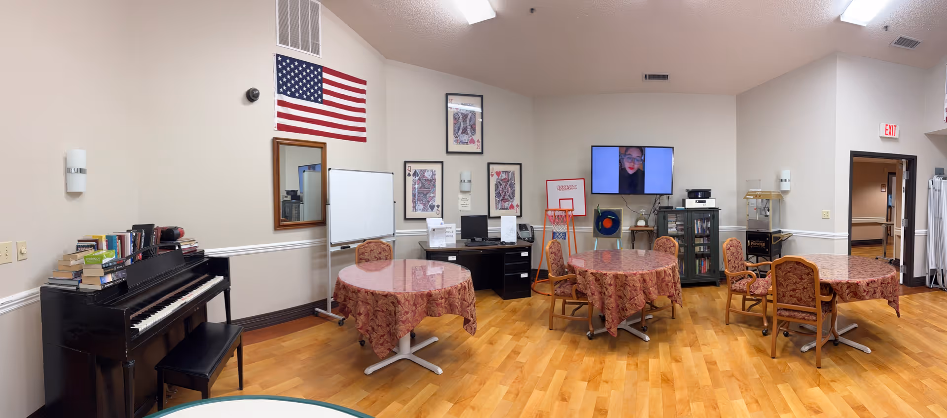 Communal senior living room with round tables covered in patterned tablecloths, chairs, a piano, wall-mounted TV, and an American flag on a hardwood floor.