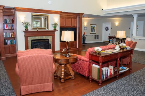 Comfortable lounge with upholstered chairs and sofas arranged around a fireplace, built-in bookshelves, lamps, and side tables.