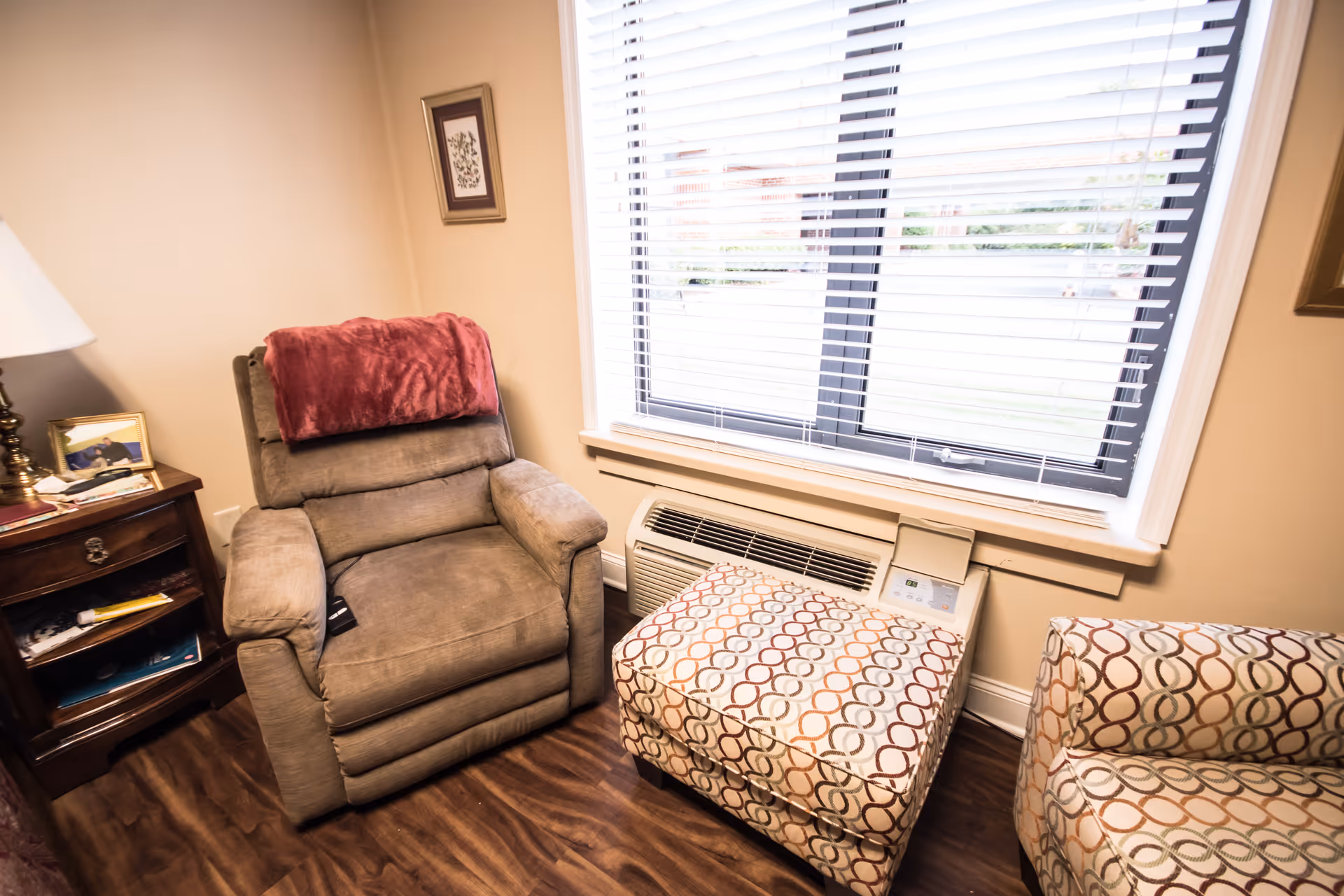 A cozy corner of a room featuring a cushioned recliner chair with a red throw draped over the back, a patterned ottoman, and a matching patterned chair. Next to the recliner is a wooden side table with a lamp, a framed photo, and some papers. A window with white blinds lets in natural light, and below the window is a heating and air conditioning unit. The floor is wood, and the walls are painted beige with a small framed picture hanging.