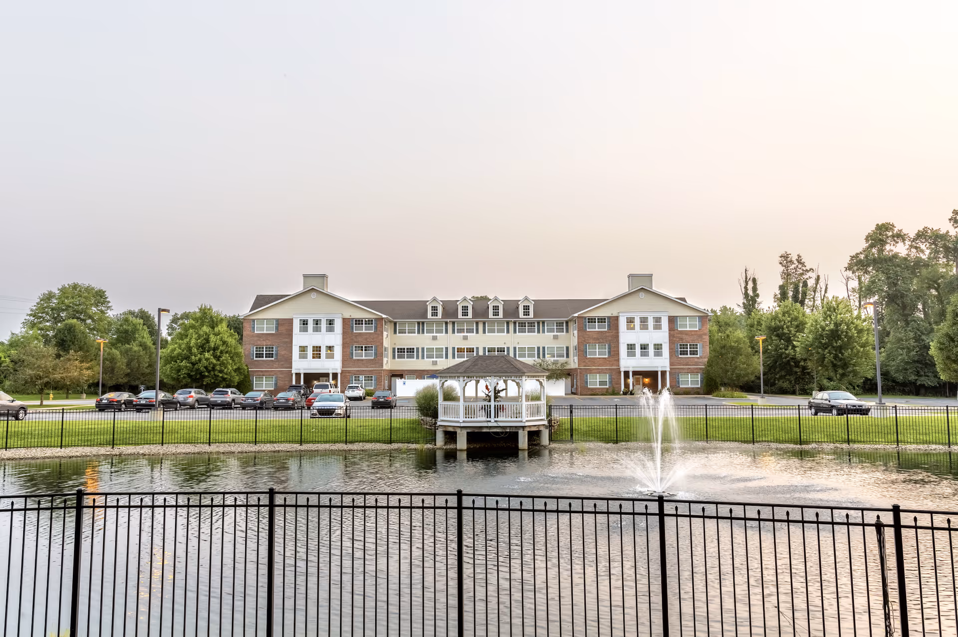 A three-story senior living facility building with a brick and beige exterior, viewed from across a pond with a water fountain and a white gazebo. There is a black metal fence surrounding the pond, and several cars are parked in front of the building. Trees and greenery surround the area under a clear sky.