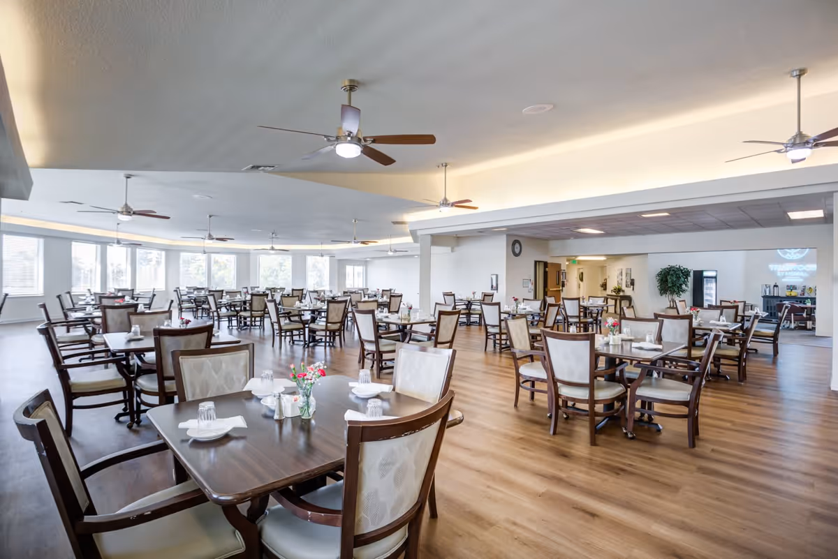 Spacious dining room with multiple wooden tables and cushioned chairs arranged neatly. Each table is set with glasses, napkins, and small flower vases. The room has large windows allowing natural light to fill the space, ceiling fans, and wood flooring.