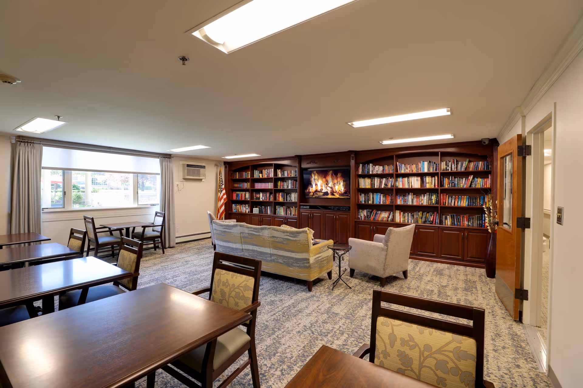 A bright community lounge with tables and chairs, sofas and an armchair facing a built-in bookshelf and fireplace display.