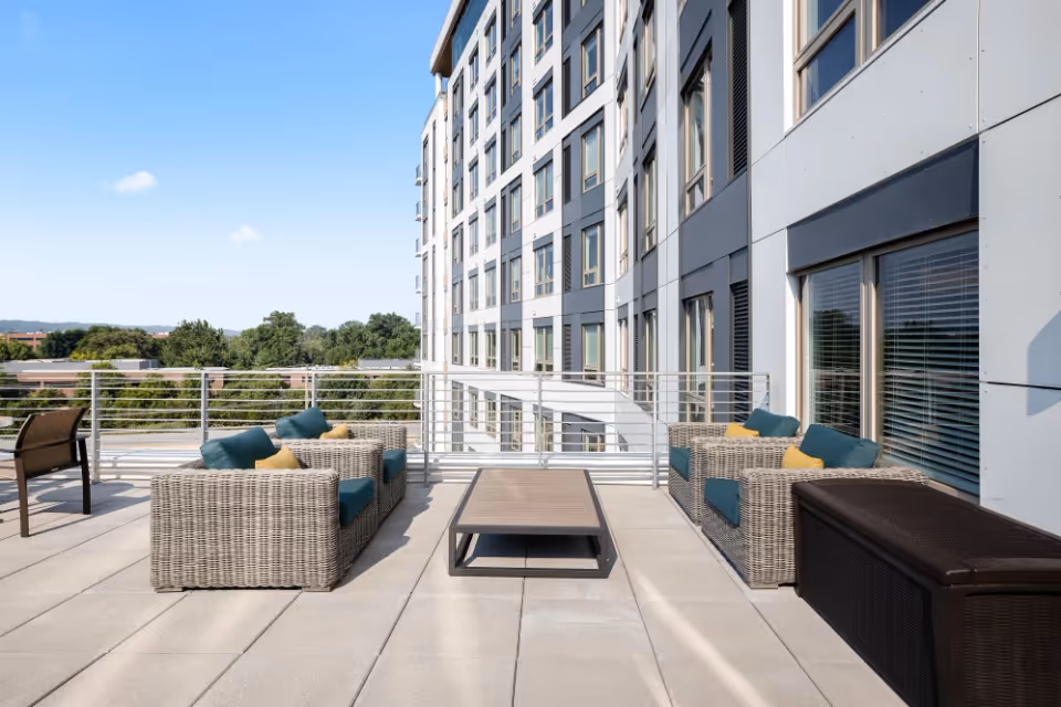 Outdoor patio area at Revelle King of Prussia featuring two wicker sofas with blue and yellow cushions, a rectangular coffee table, and a brown storage bench. The patio overlooks trees and other buildings under a clear blue sky.