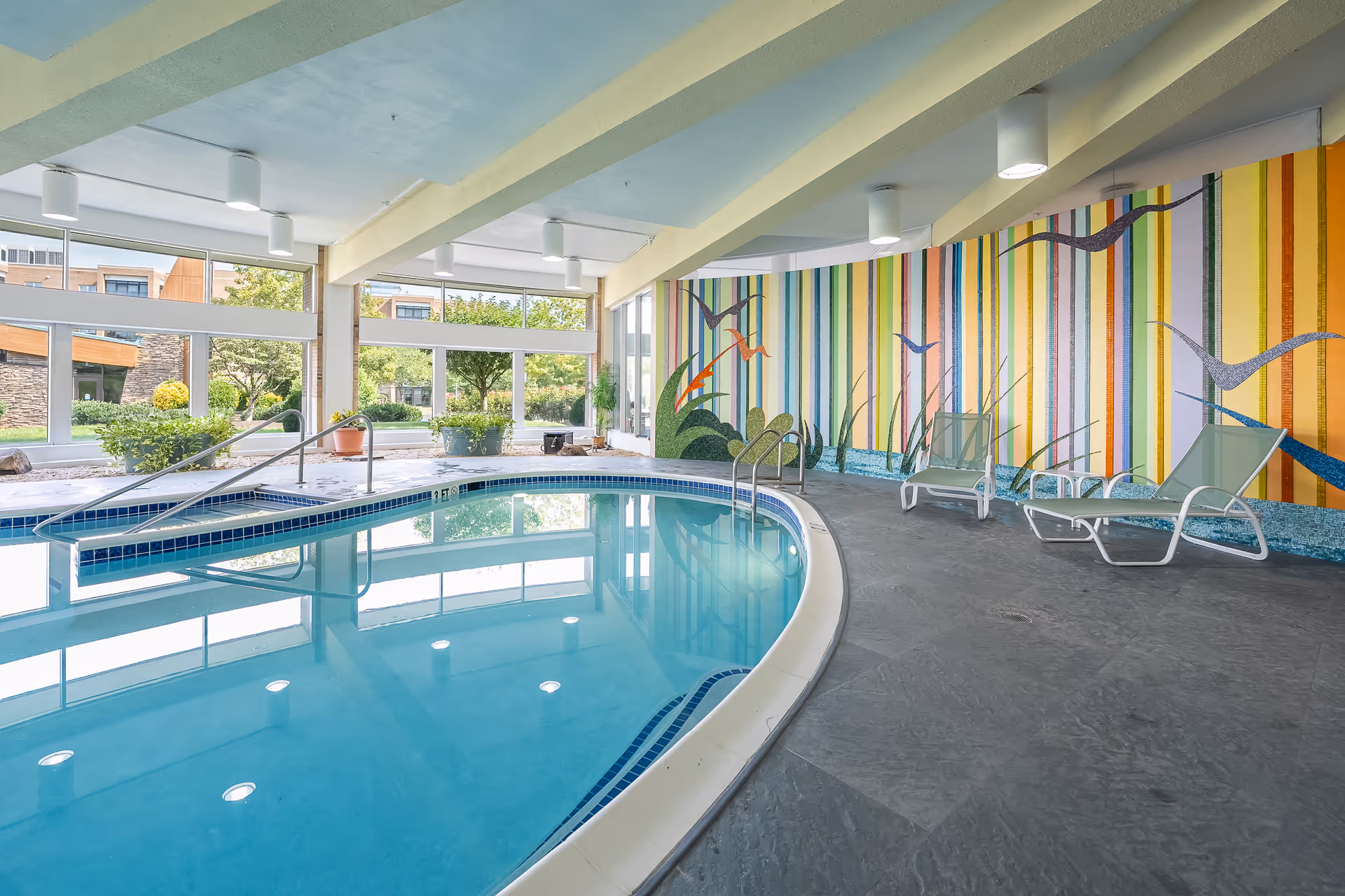 Indoor swimming pool area with clear blue water, surrounded by large windows showing outdoor greenery. The pool area has a colorful mural with vertical stripes and bird designs on one wall, and several lounge chairs placed along the poolside.