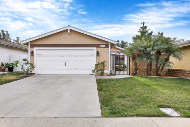Front exterior view of a single-story house with a beige facade, a white double garage door, a concrete driveway, and a green lawn with some plants and trees on the right side. The sky is partly cloudy.
