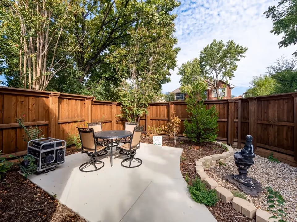 Outdoor patio area with a round metal table and four chairs on a concrete slab, surrounded by a wooden fence and various trees and plants. There is a small black statue on a pedestal in a gravel bed bordered by bricks, and a portable generator is placed near the fence.
