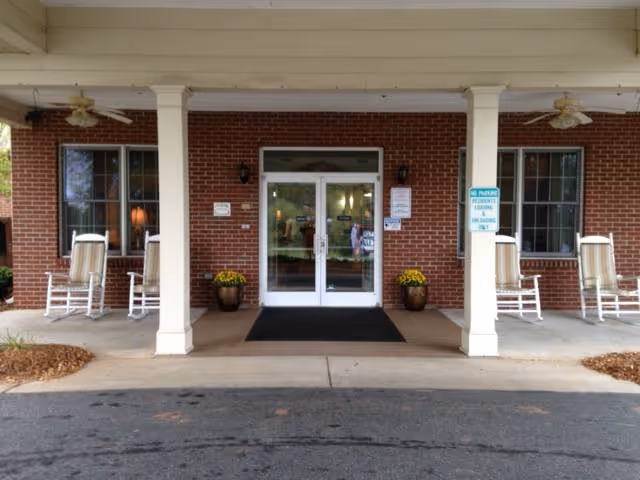 Entrance of a senior living community building with a covered porch supported by white columns. There are four white rocking chairs with cushions placed on the porch, two on each side of the double glass doors. Two potted plants with yellow flowers flank the entrance. The building exterior is made of red brick. There are signs posted near the entrance and a black mat in front of the doors.