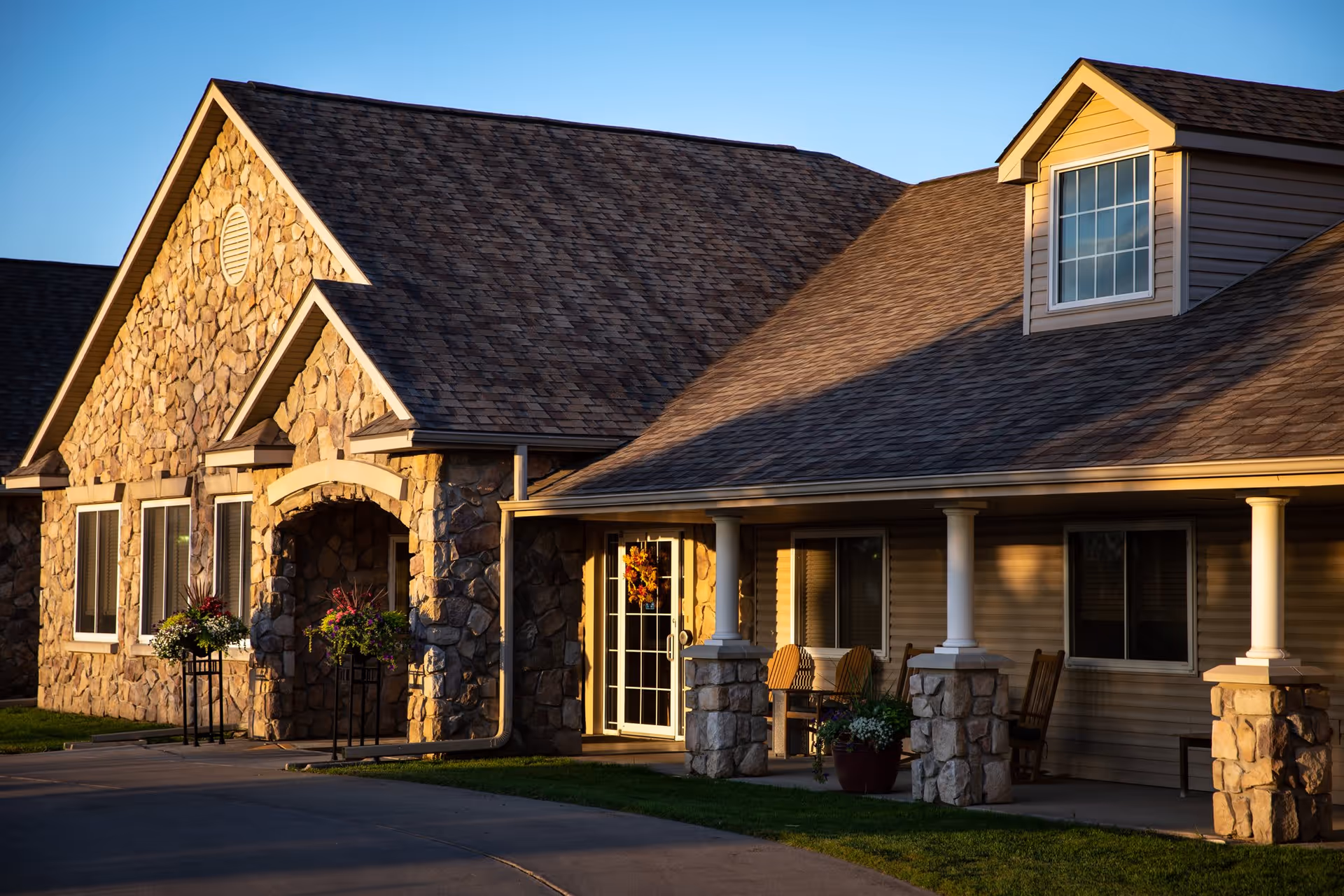 Exterior view of a stone and siding building with a porch supported by stone pillars, featuring rocking chairs and potted plants, illuminated by warm sunlight.