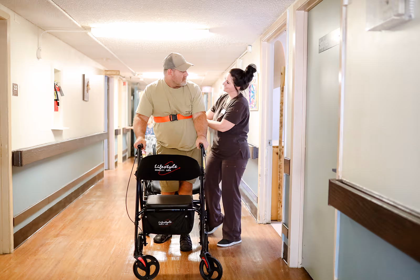 A man using a walker is assisted by a female caregiver in a well-lit hallway of a healthcare facility. The man is wearing a beige shirt and cap, and the caregiver is smiling and supporting him. The hallway has wooden floors, handrails, and several doors along the walls.