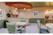 A cozy common area in a senior living facility featuring round tables with green cushioned chairs, a reception desk with a flower arrangement, framed artwork on the walls, and a computer workstation in the corner. The ceiling has recessed lighting and pendant lights above the reception desk.
