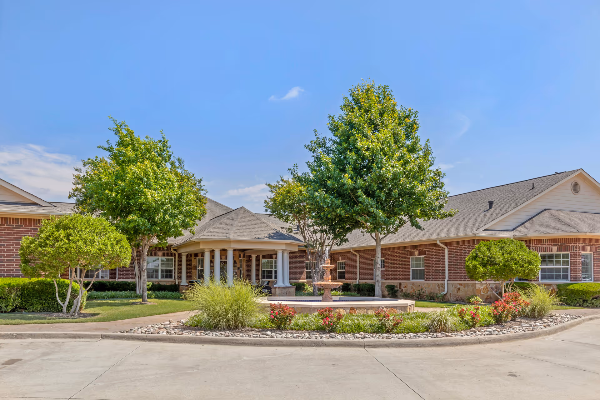 Exterior view of a single-story brick building with a covered entrance supported by columns, surrounded by neatly trimmed trees, bushes, and a landscaped area with a fountain in front under a clear blue sky.