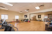 Large bright multipurpose activity room with exercise machines, tables and chairs, a wall-mounted TV, and a kitchenette area.