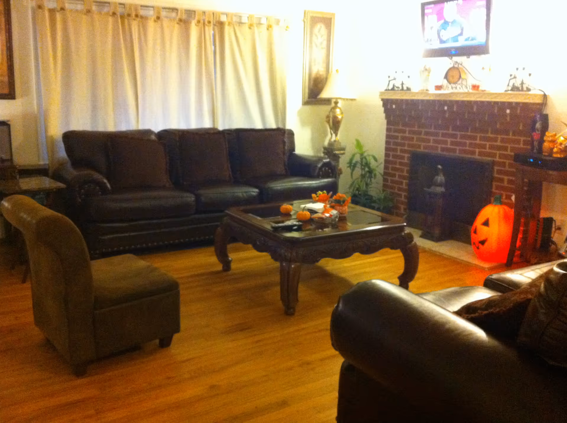 A cozy living room with a dark brown leather sofa, a matching armchair, and a wooden coffee table decorated with small pumpkins and fall-themed items. There is a brick fireplace with a large orange jack-o'-lantern decoration on the floor next to it. A flat-screen TV is mounted above the fireplace, and a lamp and framed artwork are on the walls. The floor is wooden, and cream-colored curtains cover the window.