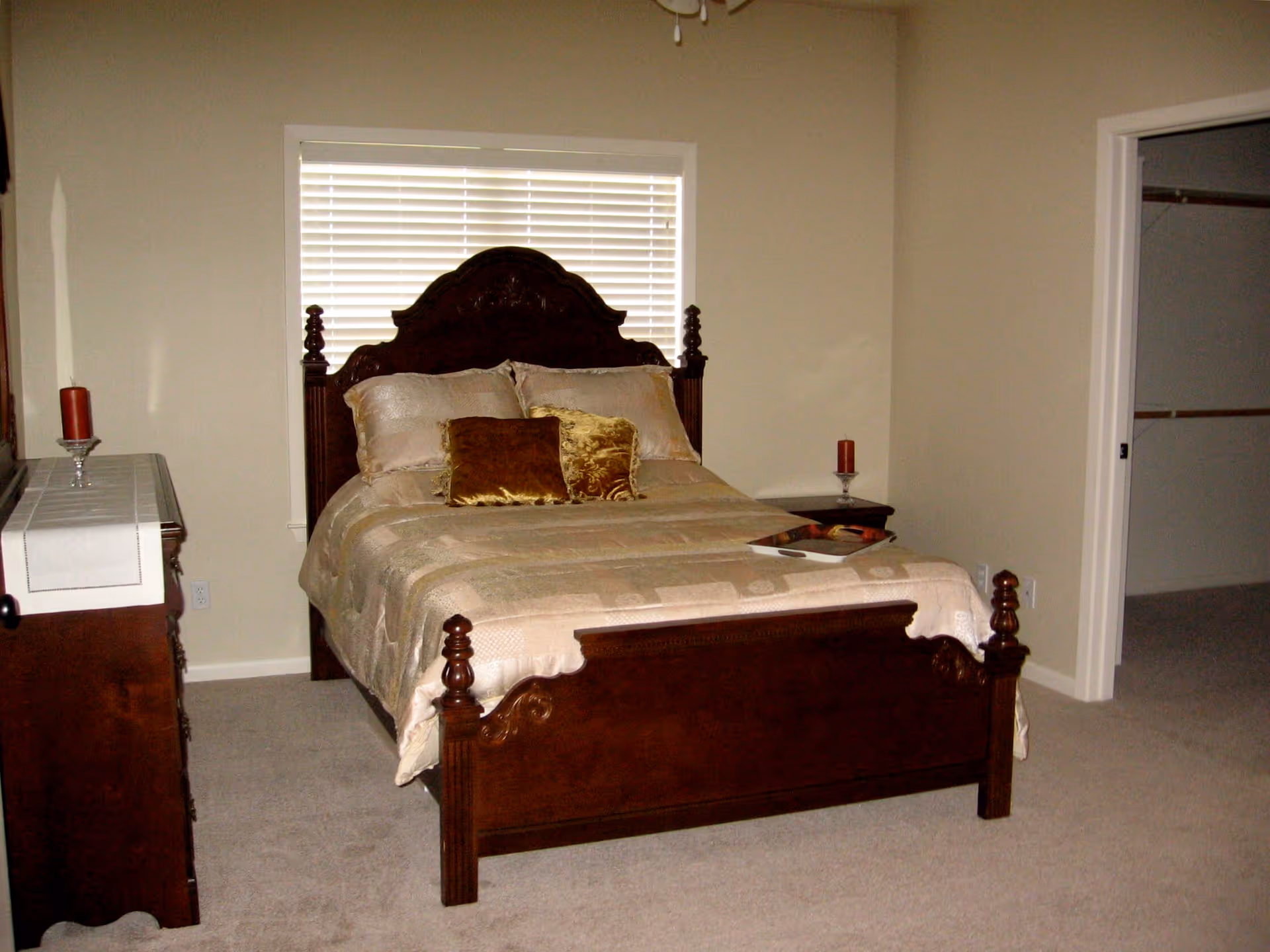 A bedroom with a wooden bed frame featuring an ornate headboard and footboard. The bed is made with beige and gold bedding and several pillows. There is a wooden dresser with a white runner and a red candle on top to the left of the bed. A small round wooden nightstand with a red candle is on the right side of the bed. The room has beige walls, a window with white blinds behind the bed, and carpeted flooring. An open doorway leads to another room.