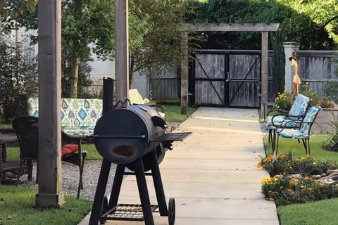 Outdoor garden area with a black barbecue grill on wheels in the foreground, a paved walkway leading to a wooden gate, benches with colorful cushions on either side, green grass, flowers, and trees providing shade.