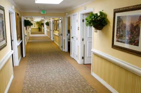 A long carpeted hallway in a senior living facility with framed art and hanging plants.