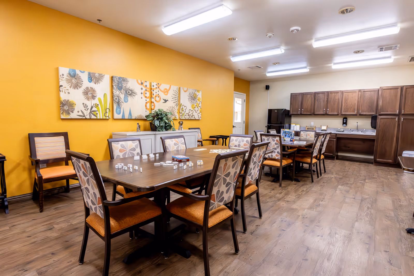 A spacious dining room with two wooden tables surrounded by chairs with patterned backs and orange seats. The room has a yellow accent wall decorated with three floral paintings and a sideboard with a plant. The opposite wall features a kitchenette area with dark wooden cabinets, a black refrigerator, and a sink. The floor is wooden, and the ceiling has fluorescent lighting.