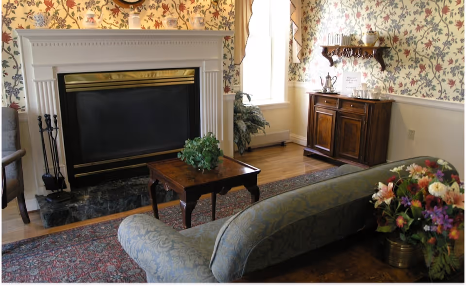 A cozy living room with a white fireplace featuring gold accents, a small wooden coffee table with a green plant, a patterned area rug, a floral upholstered sofa, and a wooden cabinet with a silver tea set and glasses on top. The walls have floral wallpaper and there is a window letting in natural light.