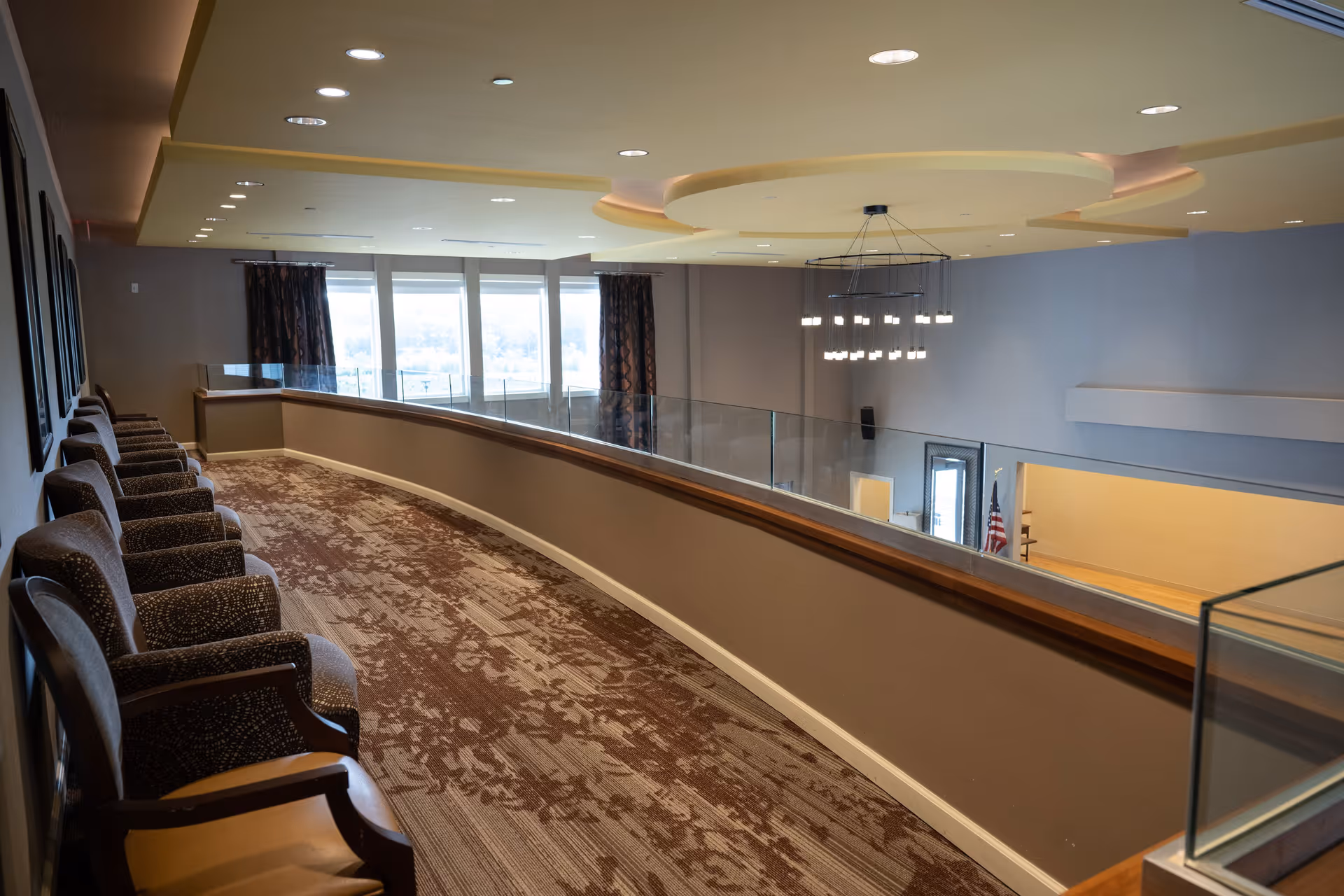 A curved indoor balcony area with a row of upholstered chairs along the left side. The space features a patterned carpet, glass railing, and a modern chandelier hanging from the ceiling. Large windows with curtains allow natural light to enter the space.