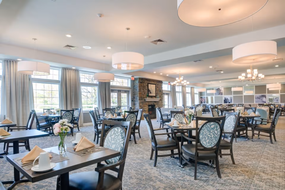 A spacious and well-lit dining room with multiple tables set with napkins, cups, and flowers. The room features large windows with curtains, modern chandeliers, and a stone fireplace in the background. The chairs have patterned upholstery and the overall decor is elegant and inviting.