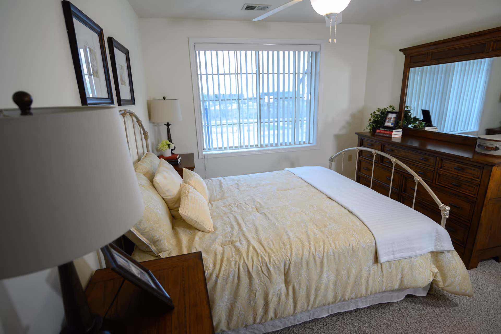 Bright furnished bedroom with a bed, wooden dresser and a window with vertical blinds.