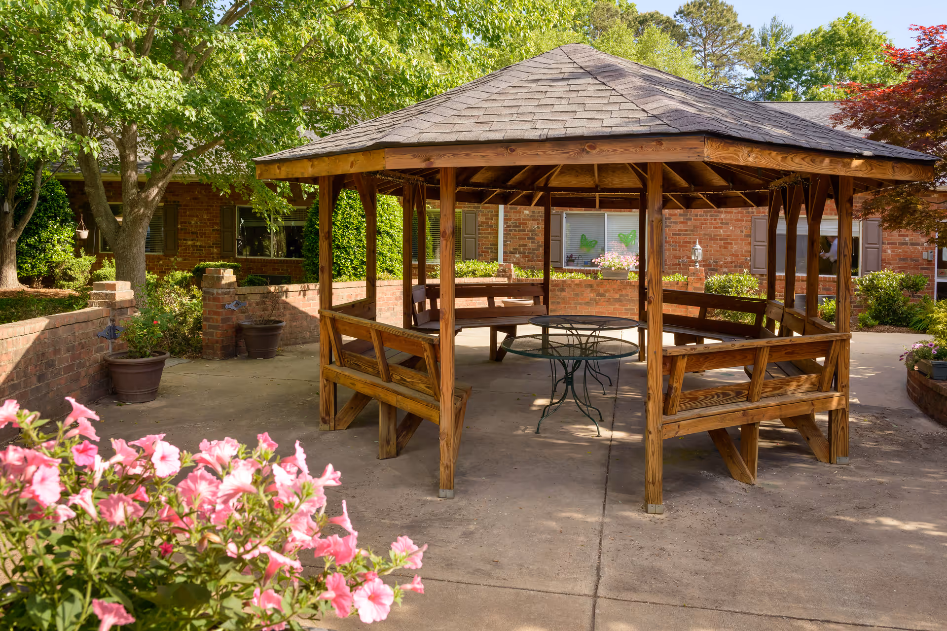A wooden gazebo with a shingled roof and built-in benches surrounding a round metal table, situated on a concrete patio. The area is surrounded by brick walls, potted plants, green trees, and flowering bushes, with a brick building in the background.