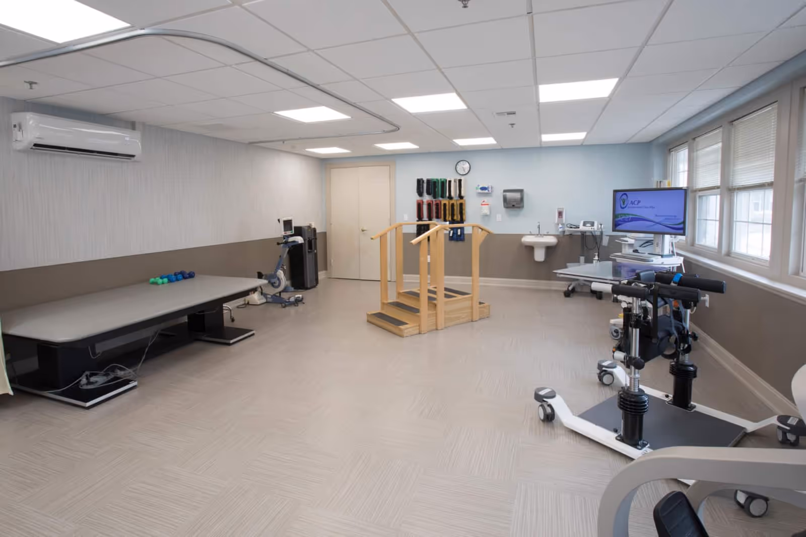 A physical therapy room with exercise equipment including a therapy table with small weights, a stationary bike, parallel bars for walking exercises, and other rehabilitation devices. The room has large windows on one side, a wall-mounted air conditioner, a sink, and a clock on the wall.