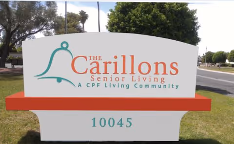 Outdoor sign for The Carillons Senior Living, a CPF Living Community, with the address number 10045 displayed below the community name. The sign is white with teal and orange text, set on a red base, with trees and a road visible in the background.