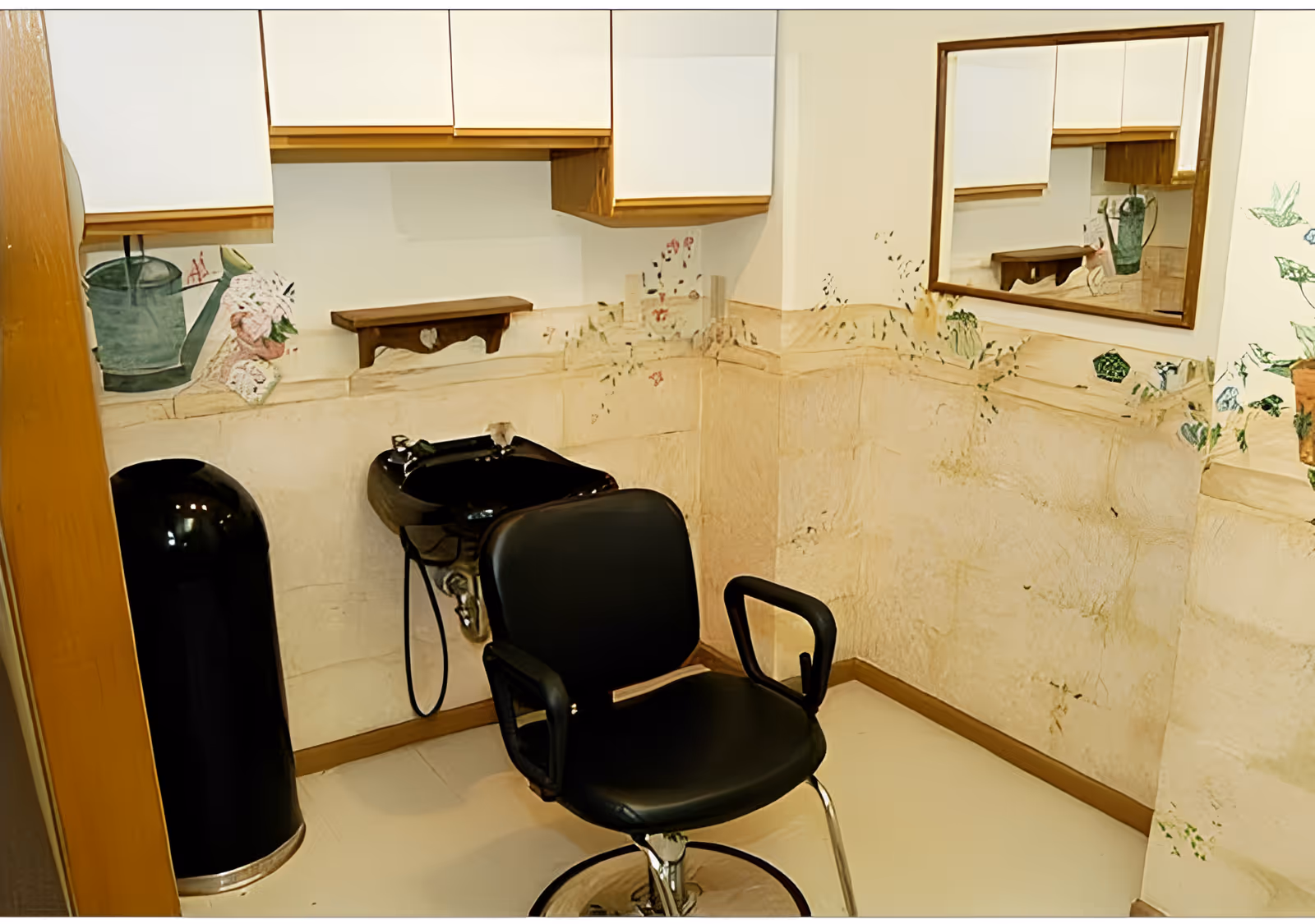 A small hair washing station with a black salon chair and a black sink basin attached to the wall. The walls have a decorative mural with plants and flowers, and there is a mirror on the right wall reflecting part of the room. White cabinets are mounted above the sink area.