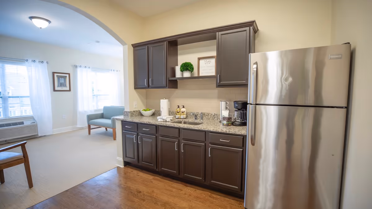 A kitchenette area with dark brown cabinets, a granite countertop, a stainless steel refrigerator, a coffee maker, and a small sink. In the background, there is a living room area with a light blue loveseat, a wooden chair, two windows with white curtains, and a framed picture on the wall.