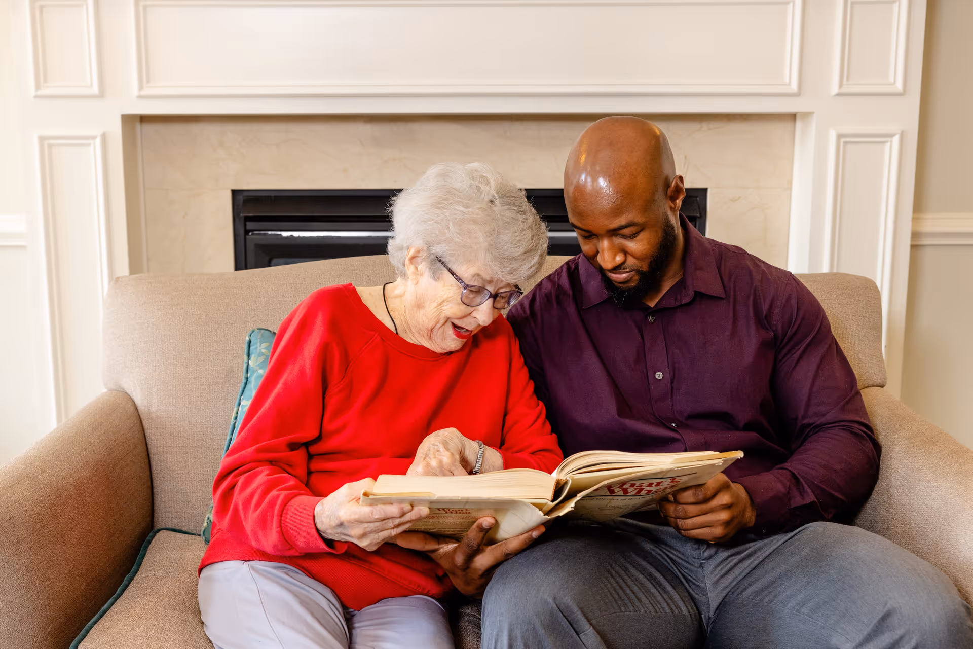 An elderly woman in a red sweater and a man in a dark purple shirt sit closely together on a beige couch, looking at and reading a book. They are in a cozy room with a fireplace in the background.