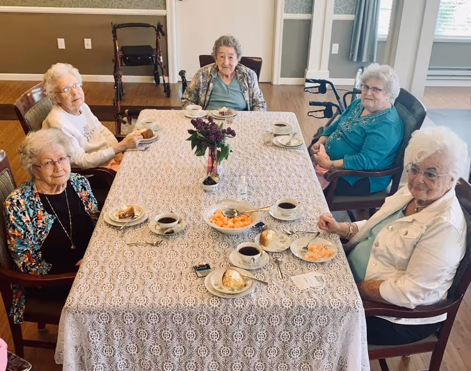 Five elderly women sitting around a rectangular table covered with a lace tablecloth, enjoying coffee and snacks in a well-lit room with wooden flooring. There is a vase with purple flowers in the center of the table, and a walker is visible in the background near the wall.