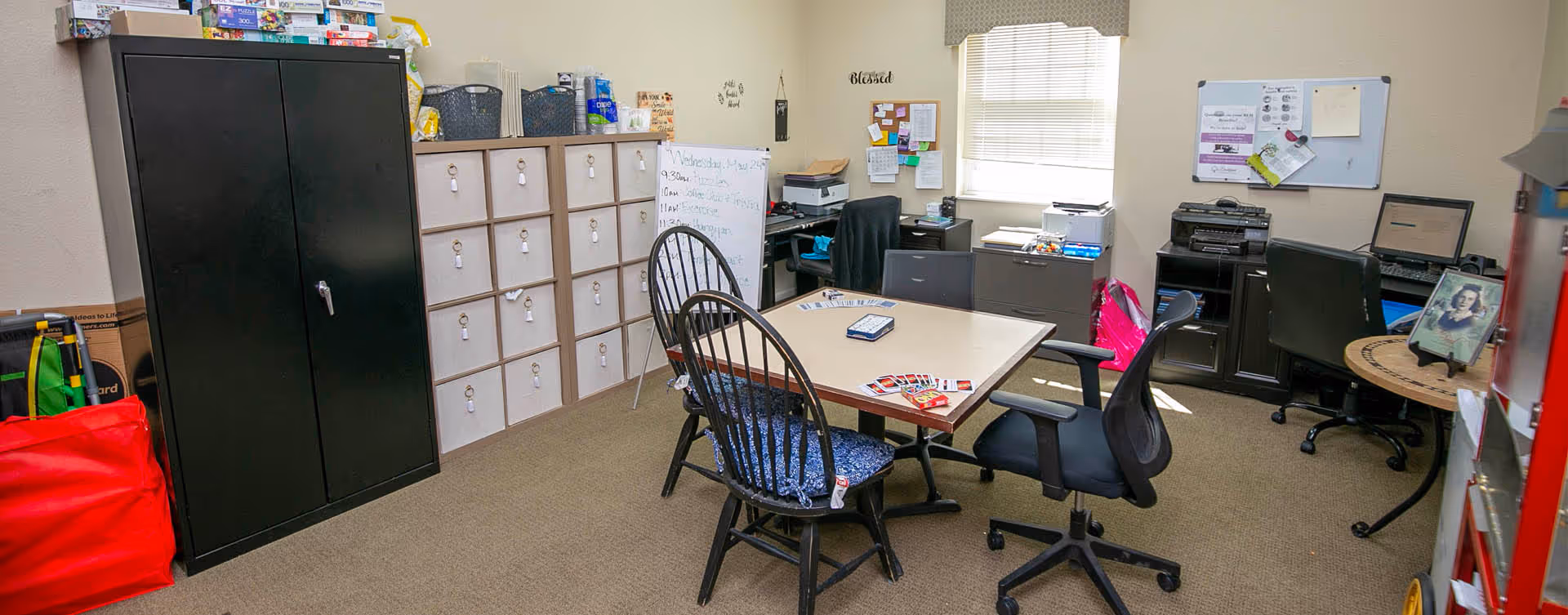 An office or activity room with a table in the center surrounded by four chairs, two black wooden chairs with blue cushions and two black office chairs. On the table are playing cards and a small box. The room has a black metal cabinet, a storage unit with multiple cubby holes, a whiteboard with a schedule, desks with computers, printers, and office supplies. A window with blinds lets in natural light, and various papers and decorations are pinned on the walls.