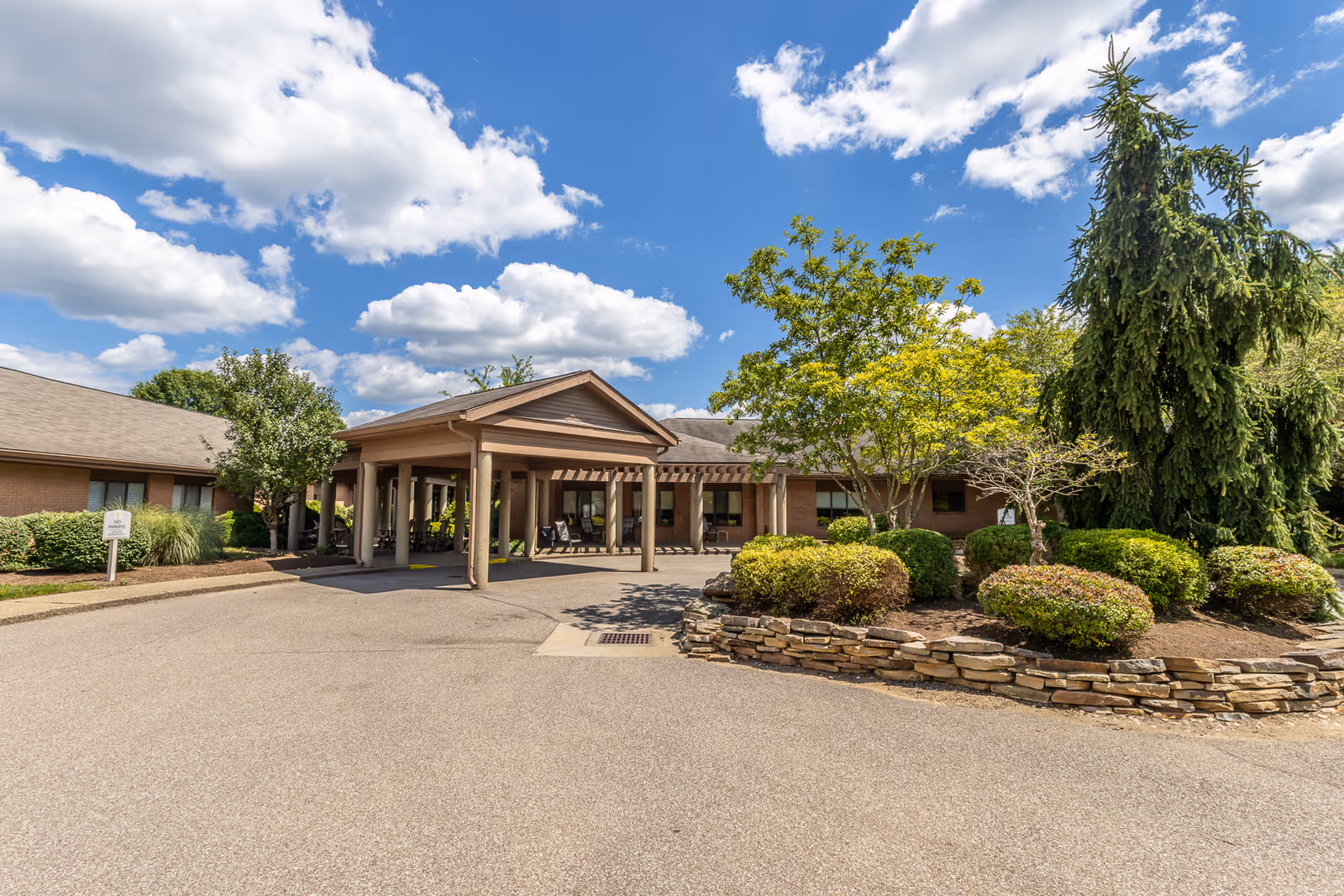 Exterior view of Elison Assisted Living & Memory Care of Marietta building entrance with a covered driveway, surrounded by landscaped bushes, trees, and a stone border under a partly cloudy blue sky.