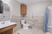 A clean, accessible bathroom featuring a toilet with grab bars on both sides, a sink with a mirror above it, a wall-mounted soap dispenser, a wooden cabinet mounted on the wall, and a shower area with a blue shower curtain.