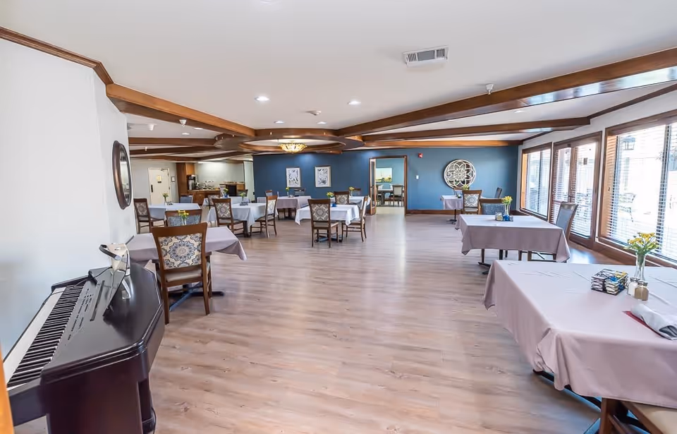 A spacious dining room with multiple tables covered in light purple tablecloths, each set with chairs and small flower vases. The room features wooden beams on the ceiling, large windows with blinds on the right side, and a piano on the left. The far wall is painted blue with decorative artwork and a large circular wall decoration.