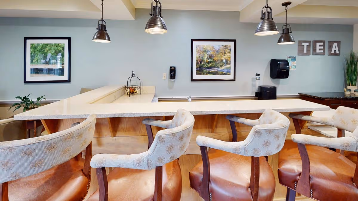 Bright common-area bar with upholstered barstools at a long countertop, pendant lights, framed artwork and a 'TEA' sign on the wall.