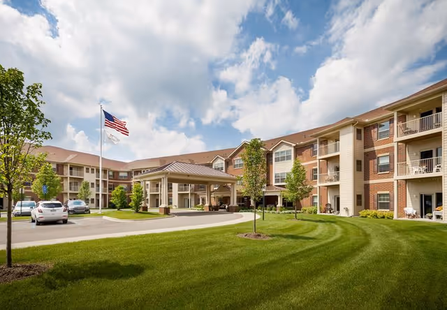Exterior view of a multi-story senior living facility with a covered entrance, well-maintained green lawn, several young trees, parked cars, and an American flag flying on a flagpole under a partly cloudy sky.