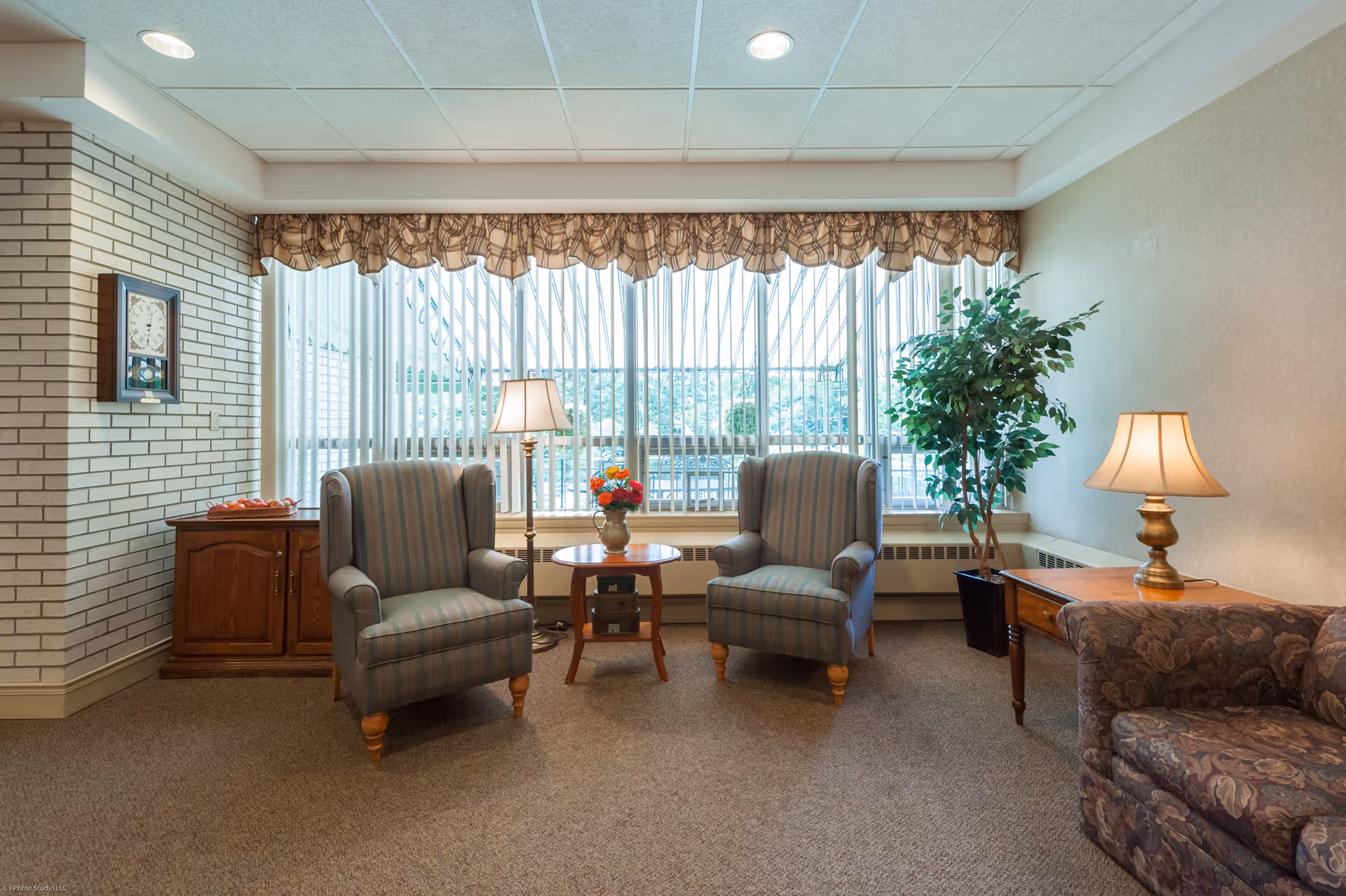 A cozy sitting area with two striped armchairs facing each other, a small round wooden table with a vase of flowers between them, a floor lamp, a potted plant, a wooden side table with a table lamp, and a floral-patterned sofa. The room has a large window with vertical blinds and a valance, beige walls, and carpeted floor.