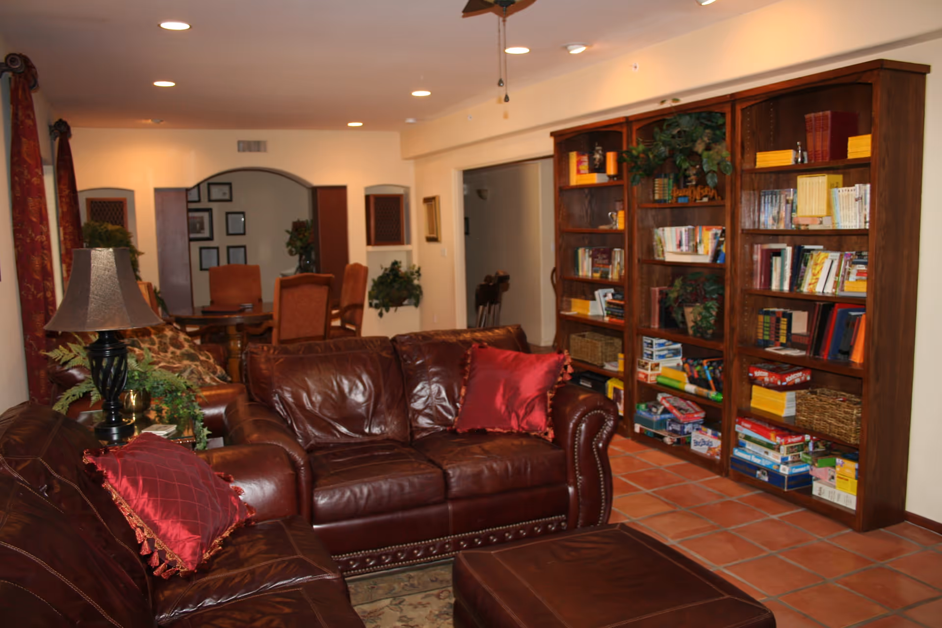 A cozy living room with brown leather sofas adorned with red pillows, a matching ottoman, a side table with a lamp and plants, and a large wooden bookshelf filled with books and board games. In the background, there is a dining area with a table and chairs, and some framed pictures on the wall.