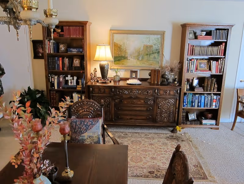 Ornate wooden sideboard and bookshelves with a lamp and painting above, viewed from a dining table with a floral centerpiece.