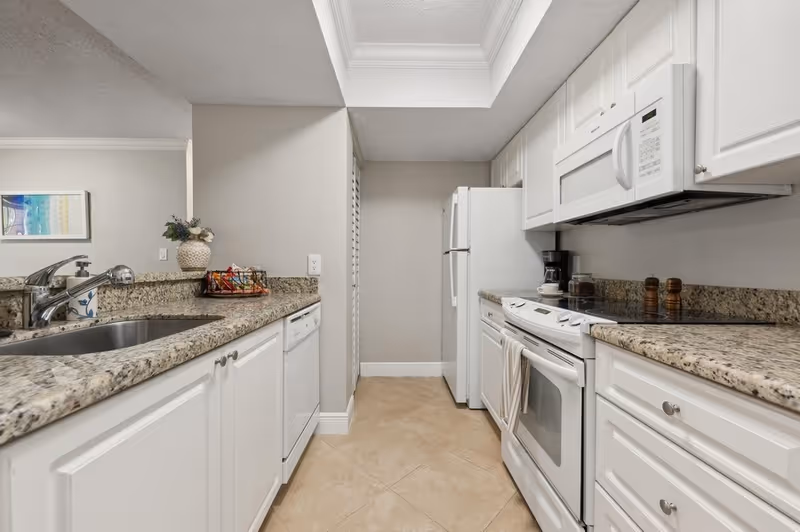 A clean, modern kitchen with white cabinets and granite countertops. The kitchen features a stainless steel sink, a white dishwasher, a white refrigerator, a white electric stove with oven, a white microwave, a coffee maker, and salt and pepper shakers on the counter. The floor is tiled with beige tiles, and there is a decorative vase with flowers and a basket with snacks on the counter.