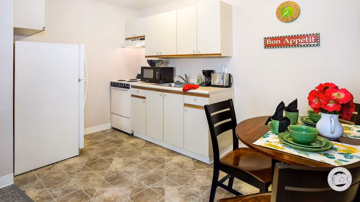 A small kitchen area with white cabinets, a white refrigerator, a stove, a microwave, a coffee maker, and a toaster. To the right, there is a round wooden dining table set with green dishes, black napkins, and a white vase with red flowers. On the wall above the table, there is a green clock and a red sign that says 'Bon Appetit'.