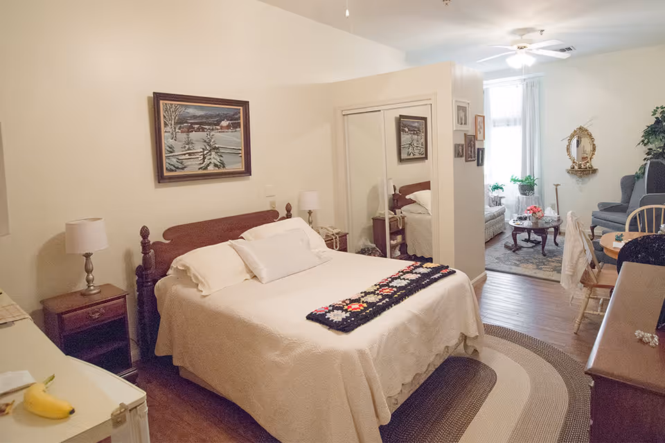 A cozy bedroom with a double bed covered in a white quilt and a colorful crocheted blanket at the foot. The room has wooden furniture including a nightstand with a lamp, a dresser, and a mirrored closet. A framed winter landscape painting hangs above the bed. The bedroom opens into a living area with a sofa, armchair, coffee table with flowers, and a wall-mounted mirror. The space is warmly lit with natural light coming through a window with white curtains.