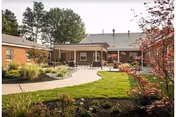 Outdoor view of a nursing center courtyard with a paved walkway, green lawn, landscaped garden beds, and a brick building in the background under a clear sky.