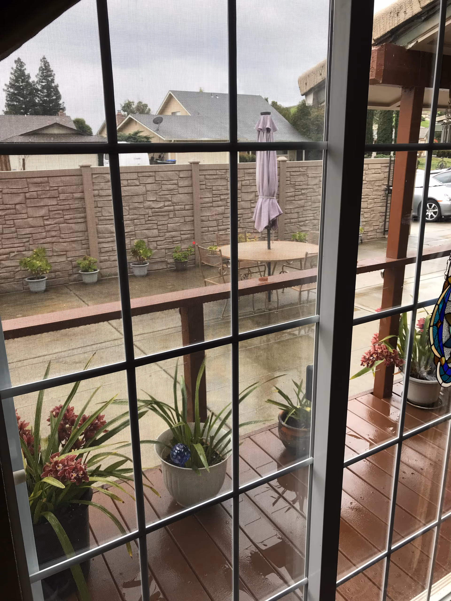 View through a glass door with grid panes showing a wet wooden deck with potted plants and flowers. Beyond the deck is a concrete patio with a round table, chairs, and a closed umbrella. A stone-patterned privacy wall and neighboring houses are visible in the background under an overcast sky.