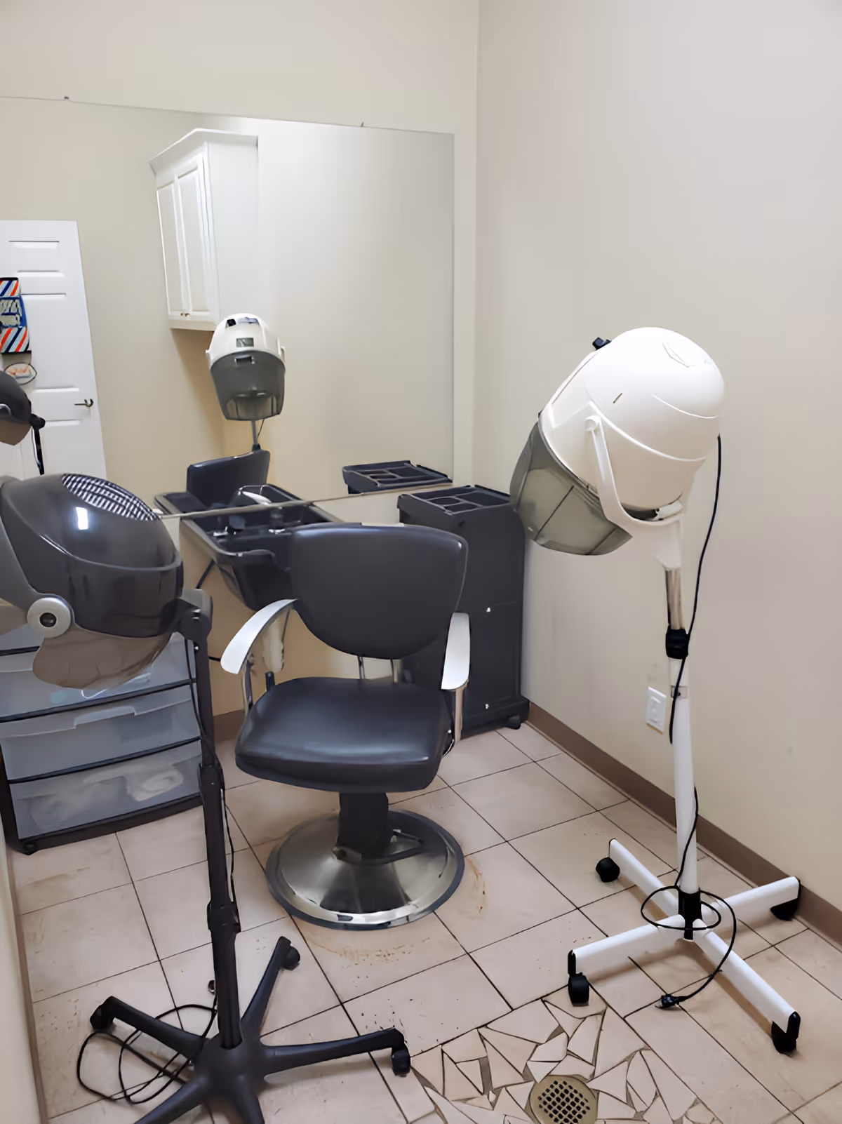 A small hair salon room with a styling chair, hooded hair dryers, mirror, and storage cart.